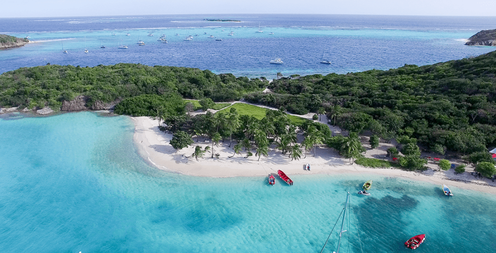 Aerial view of a tropical beach with turquoise water, lush green trees, and several boats anchored offshore.