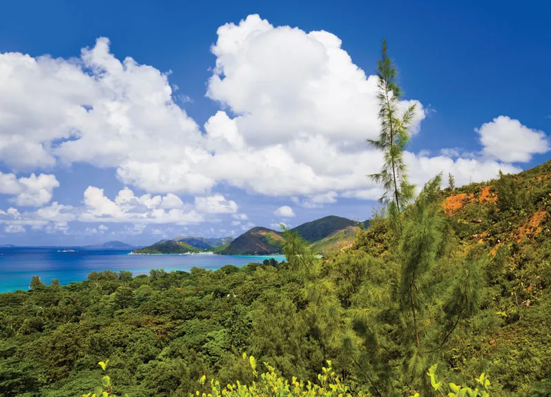 scenic view of Praslin Island, the second-largest island in the Seychelles archipelago. The landscape features lush, green, hilly terrain covered in dense tropical vegetation, with a clear turquoise sea and smaller islands visible in the distance. The sky is a vibrant blue with large, white cumulus clouds