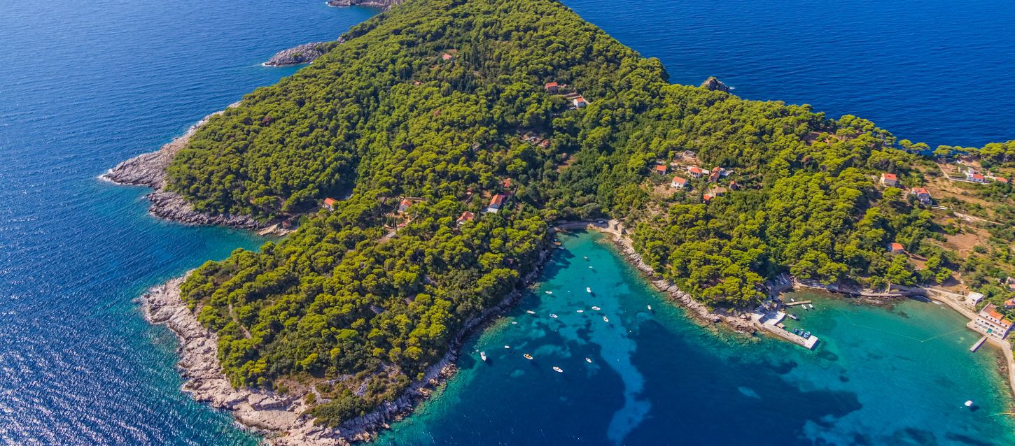 lush, green island with a rocky coastline surrounded by clear, turquoise water. Several small houses are nestled among the dense trees. On the right side of the image, a bay with calm, clear water is visible, where several yachts are anchored