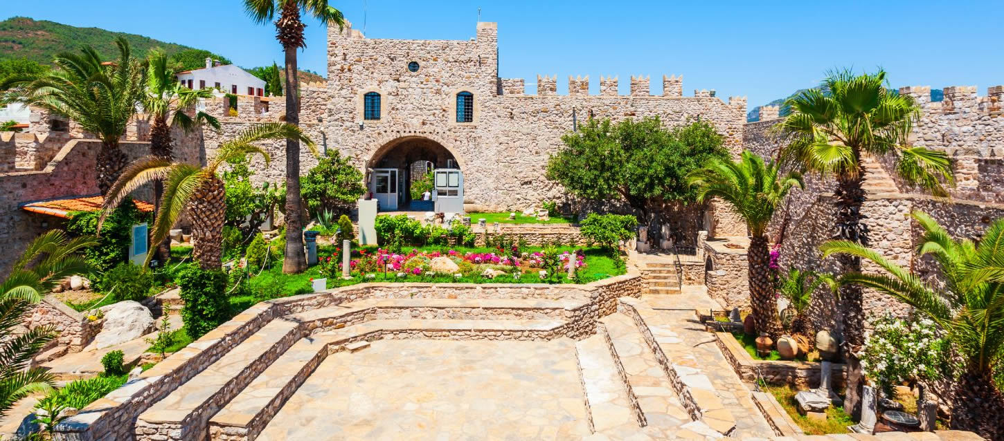The image depicts the castle's inner courtyard, which features a well-maintained garden with colorful flowers and palm trees. The castle itself is a stone structure with crenellated walls and an arched entrance leading to the interior. Stone stairs lead down from the entrance to a lower, paved area in the foreground.