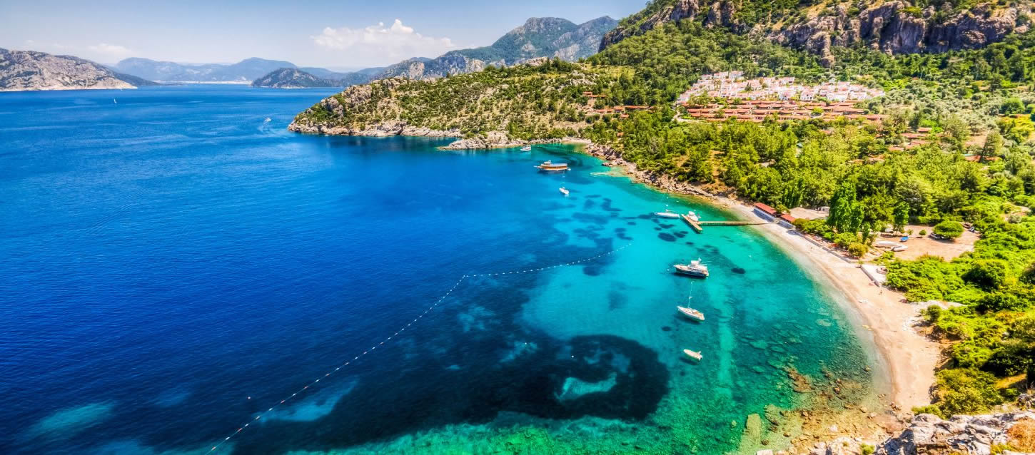 The image shows a beautiful coastal scene with a bay featuring a small sandy beach and extremely clear, turquoise-colored water. Several boats are anchored in the bay. Lush green hills and mountains covered in dense vegetation surround the bay and extend into the distance, with a few buildings visible on the hillside near the shore. The scene is bright and sunny, with a blue sky and some white clouds