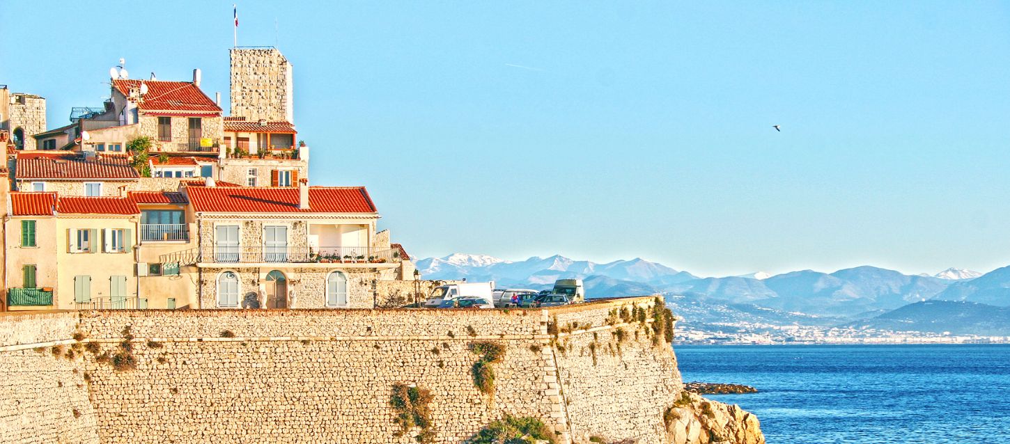 Panoramis view of Antibe's historic waterfront, featuring stone ramparts and charming Mediterranean houses with red-tiled roofs overlooking the sea and the distant mountains rising above the horizon