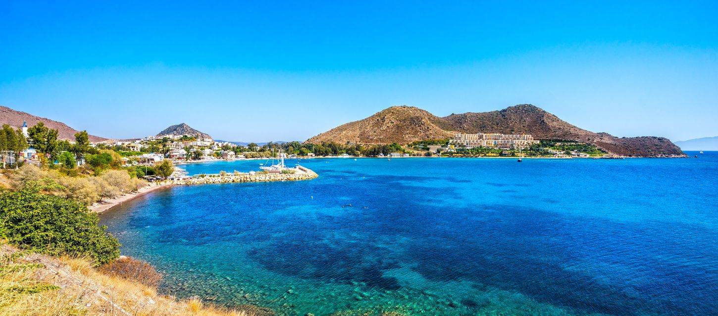 beautiful coastal landscape with a bay of clear turquoise water. In the background, there are hills and a small town with white buildings and a hotel complex. There are also a few boats anchored in the bay. The sky is bright blue and clear.