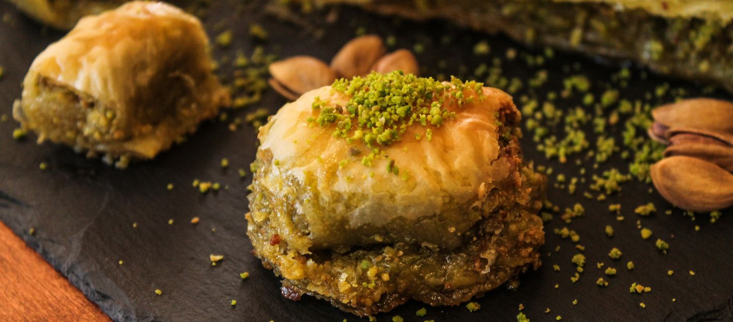 A close-up shot of two pieces of baklava, a layered pastry filled with chopped nuts and sweetened with syrup. The baklava is topped with crushed green pistachios and sits on a dark slate surface, with whole pistachios scattered around.