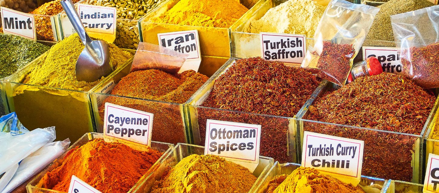 A close-up shot of a spice market stall with many different spices in open bins. The spices are vibrant shades of yellow, orange, and red. Several labels are visible, including "Mint," "Indian Curry," "Iran Saffron," "Turkish Saffron," "Cayenne Pepper," and "Ottoman Spices." A metal scoop is resting in one of the bins.