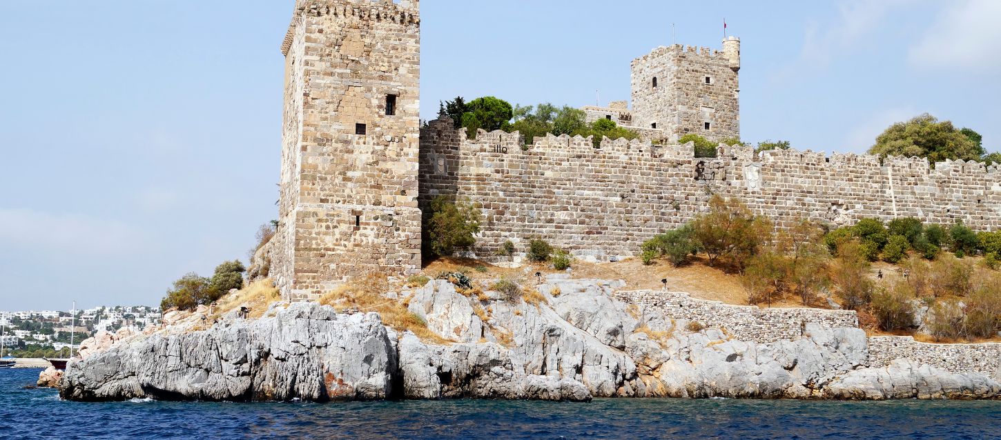 A photo of the stone walls and towers of Bodrum Castle, located on a rocky peninsula surrounded by the sea under a clear sky.