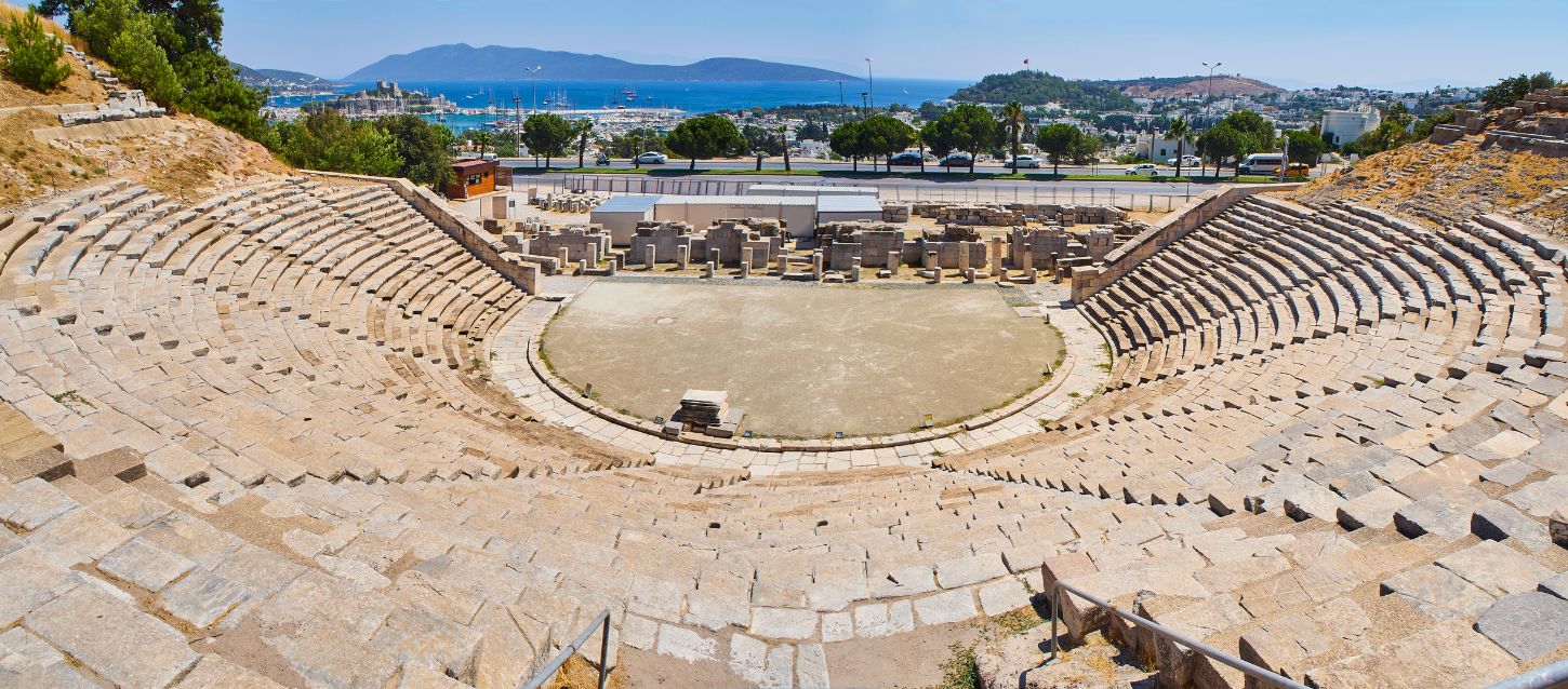 A wide-angle shot of the ancient stone amphitheater in Bodrum, Turkey, showing the tiered seating, the central stage, and the modern city and sea in the background.