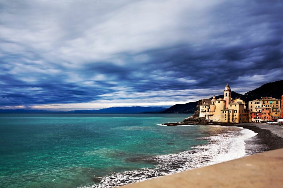 Dramatic cloudy sky over turquoise waters and the colorful coastal village of Camogli, Italian Riviera, with waves gently washing the pebbled beach.