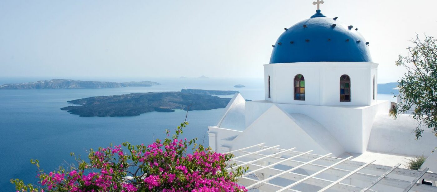 A beautiful view of a white church with a blue dome, overlooking the Aegean Sea and a volcanic caldera in Santorini, Greece. Pink bougainvillea flowers are in the foreground, adding a pop of color to the stunning landscape.