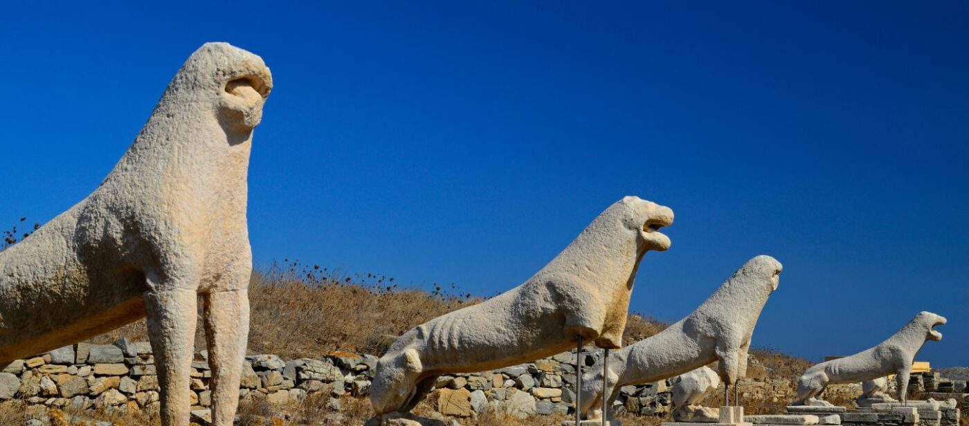 A row of ancient stone lion statues stands against a clear blue sky. These are the Terrace of the Lions on the Greek island of Delos, an archaeological site featuring a row of archaic marble lion sculptures.