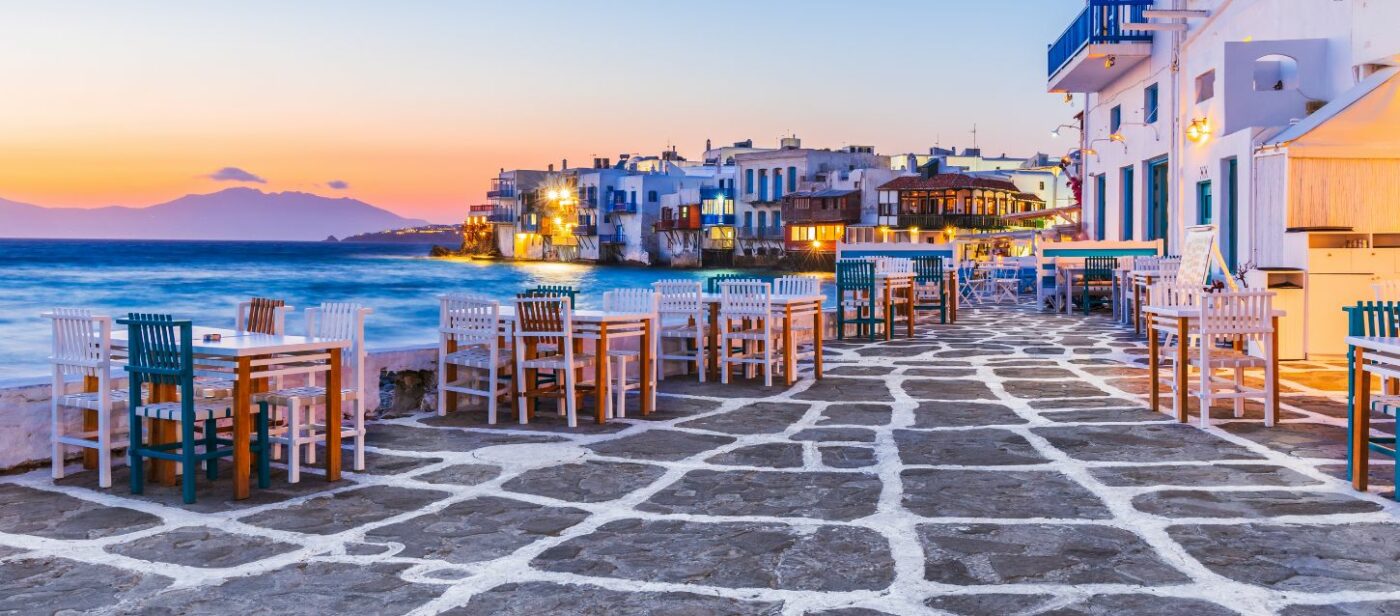 A picturesque sunset view of Mykonos Town, Greece, with traditional whitewashed buildings and waterfront restaurants lining a cobblestone street by the sea. The sky is painted with vibrant orange, pink, and purple hues, creating a serene and charming atmosphere.
