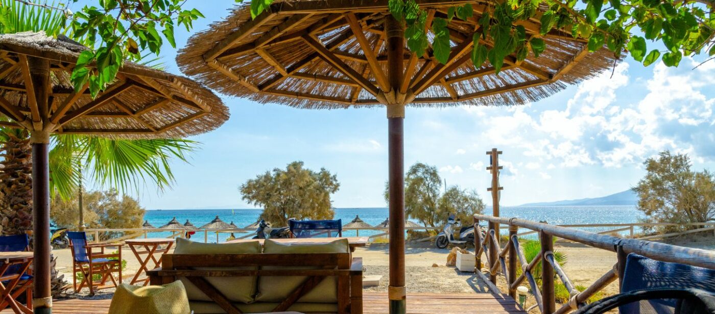 A cozy beach bar with thatched umbrellas and wooden furniture, overlooking a calm turquoise sea and a sandy shore on a sunny day.