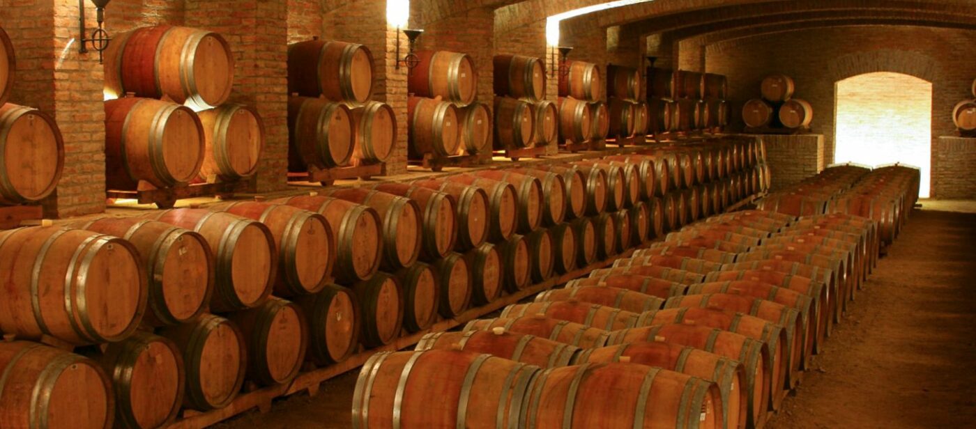 A cellar with rows of wooden barrels used for aging wine. The room has brick walls and a vaulted ceiling, creating a rustic and traditional atmosphere.