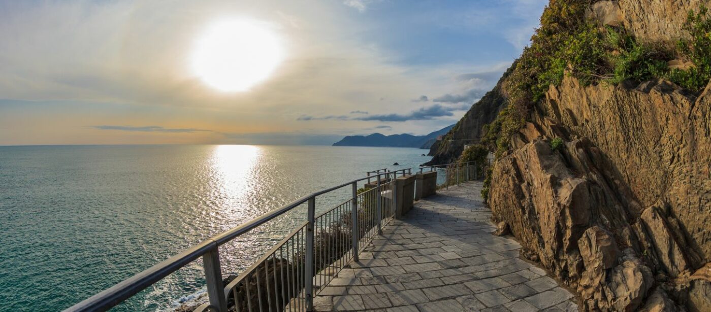 Scenic coastal walkway known as Via dell’Amore in Cinque Terre, Italy, with stone pavement, metal railing, and cliffs overlooking the calm sea at sunset.