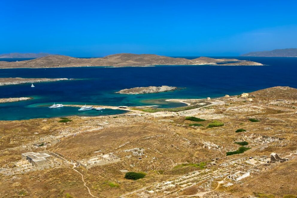 Aerial view of the ancient ruins of a city on a dry, hilly island, with the deep blue sea and other smaller islands in the background under a clear sky.