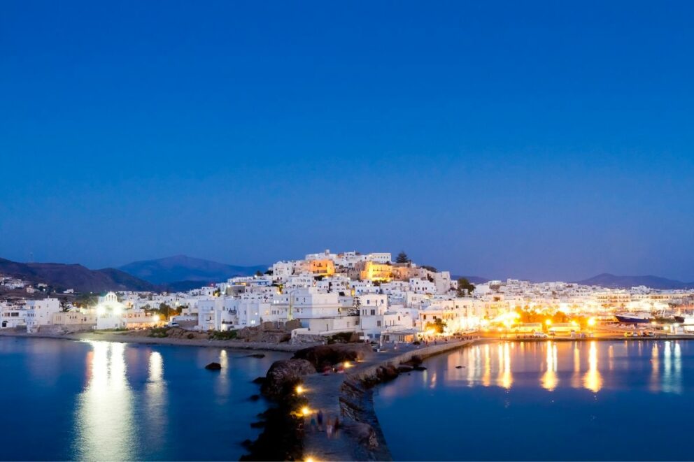 A nighttime view of a white-washed town built on a hill overlooking a harbor. A stone walkway with lights leads out into the water toward the town. The town and its reflection on the water are brightly lit. The sky is dark blue, and mountains are visible in the background. The town is Chora on Naxos island, Greece.