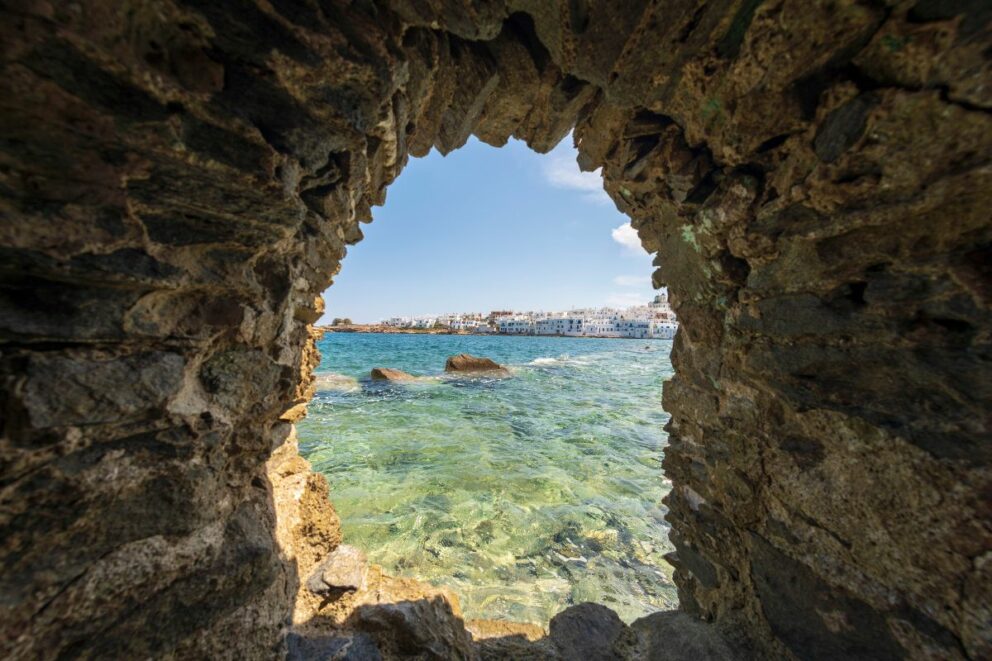 View through a rocky archway to a clear turquoise sea and a town with white buildings on a sunny day.