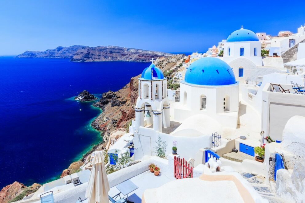 view of Santorini, Greece, an island known for its iconic white-washed buildings with vibrant blue domes and rooftops. The buildings are perched on the steep cliffs of the island's caldera, overlooking the deep blue waters of the Aegean Sea. The clear, bright blue sky complements the colors of the buildings and the sea, creating a stunning and picturesque landscape