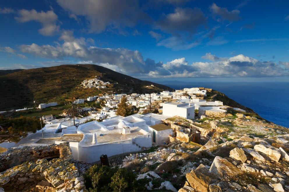 A picturesque village with traditional white buildings is perched on a rocky hill overlooking the sea under a dramatic blue sky with clouds. The village is located on the Greek island of Sikinos.