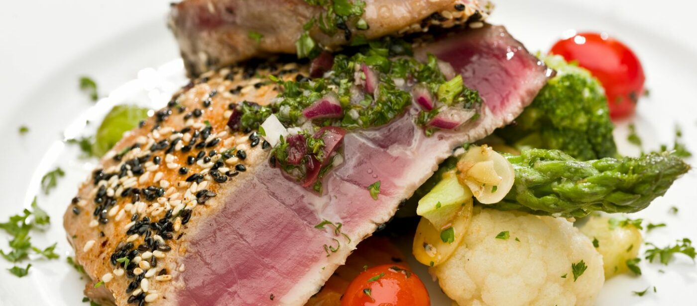 A view of the Acropolis of Lindos in Rhodes, Greece. The image shows ancient stone columns and a wall on a cliff overlooking the turquoise sea and a small bay. The sky is bright blue and clear.