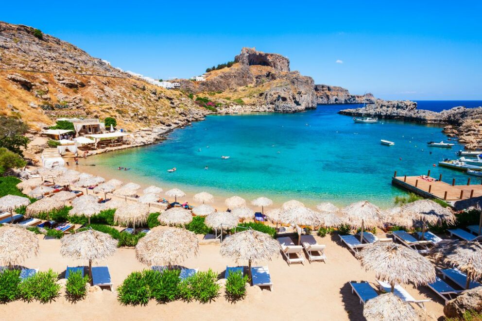 A beautiful sandy beach with straw umbrellas and sun loungers in a small, turquoise bay surrounded by rocky cliffs. The photo captures St. Paul's Bay in Lindos, Rhodes, Greece.