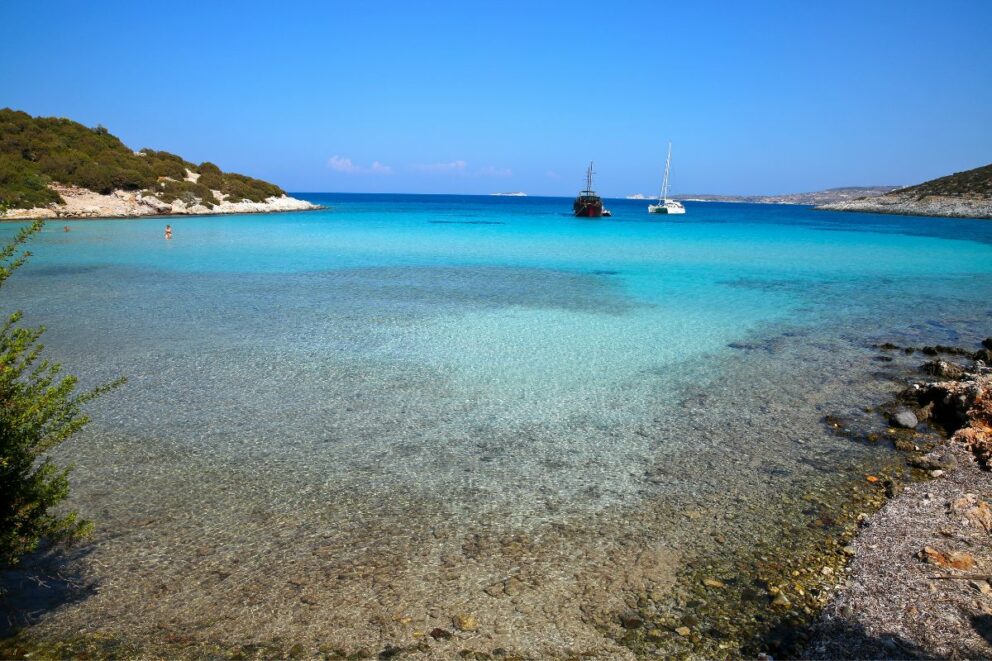 This is a photograph of a calm, clear turquoise bay. The water is so transparent that the rocky seabed is visible near the shore. In the background, two boats are anchored, and the bay is surrounded by green, rocky hills under a clear blue sky.