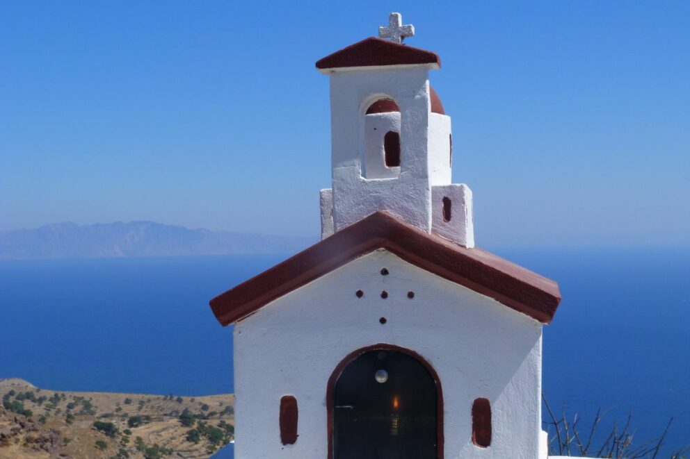 A small, white chapel with a red roof and bell tower stands on a hill overlooking a bright blue sea with a distant landmass on the horizon.