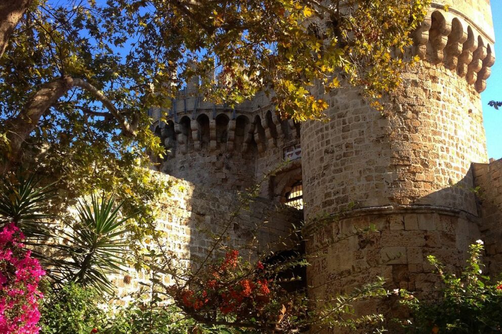 portion of the Palace of the Grand Master of the Knights of Rhodes, a medieval castle. In the foreground, there are trees with yellowing leaves and green plants with pink and red flowers, which partially obscure the stone walls of the castle. The main tower is visible on the right side of the image, with a section of the fortified wall and arched windows to its left. The sky is a clear blue.