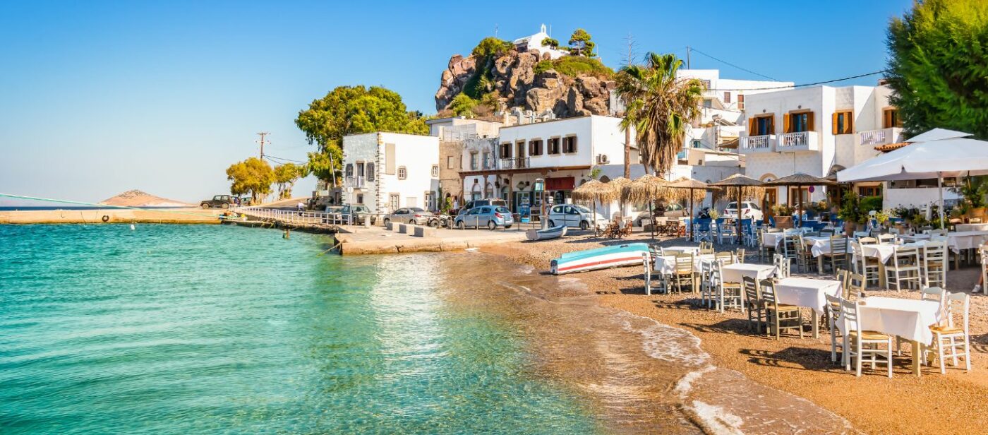 A sunny day on a beach in Patmos, Greece, with clear turquoise water and a small sandy beach. On the beach, there is a restaurant with white tables and umbrellas. The background features traditional whitewashed buildings and a rocky hill with a small chapel on top. A small boat is visible on the sand, and several cars are parked along the road next to the beach.