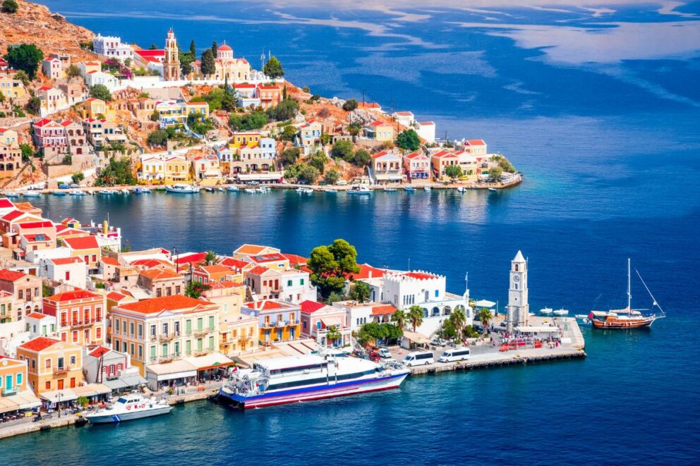 A picturesque view of the colorful houses of Symi island, Greece, built on a hillside overlooking a harbor with boats and a clock tower. The scene shows the vibrant neoclassical architecture of the town and the deep blue water of the Aegean Sea.