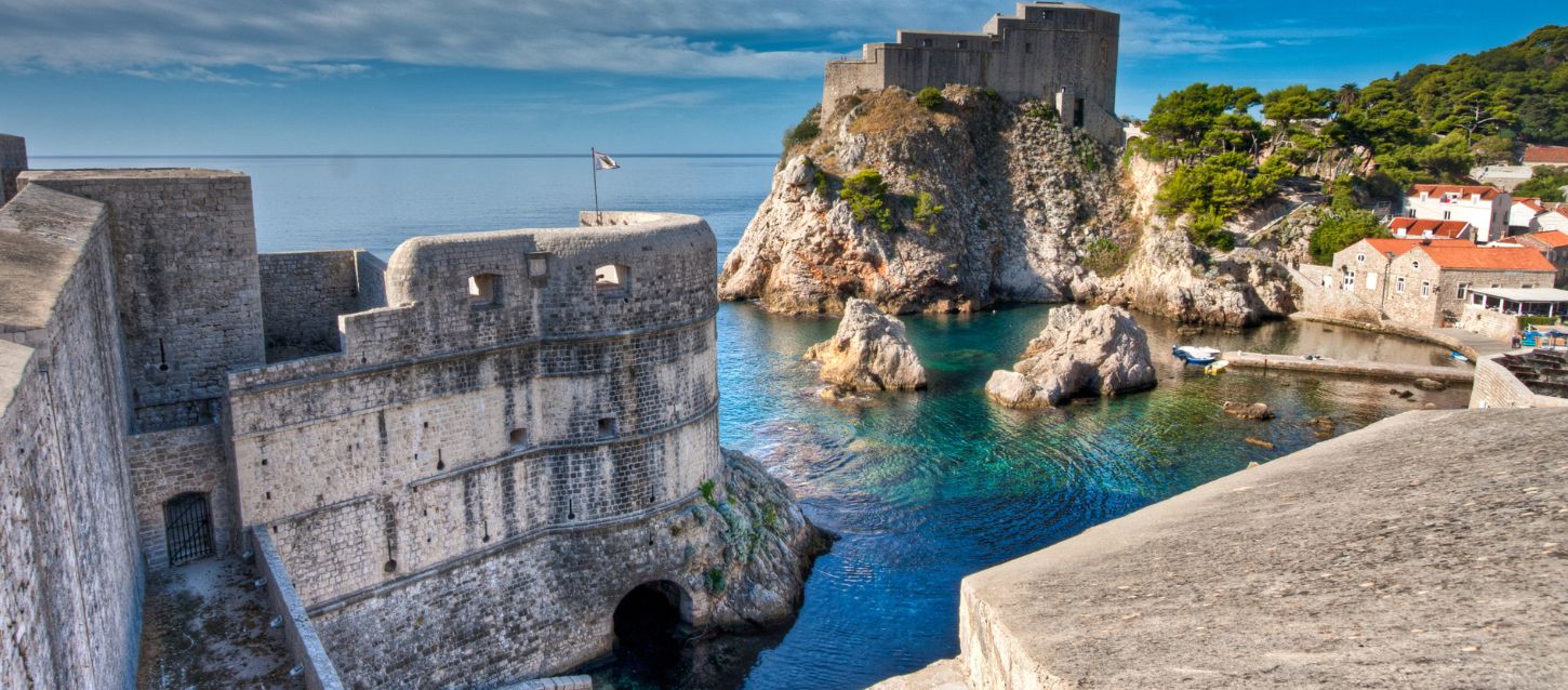 Fort Lovrijenac, in Dubrovnik, Croatia. The fortress on a large rock on the coast, overlooking a small bay with turquoise water. In the foreground, there is a large, flat, stone wall. To the right of the bay, there are several buildings with red-tiled roofs. The sky is blue with some clouds, and the surrounding area is green with trees and vegetation.