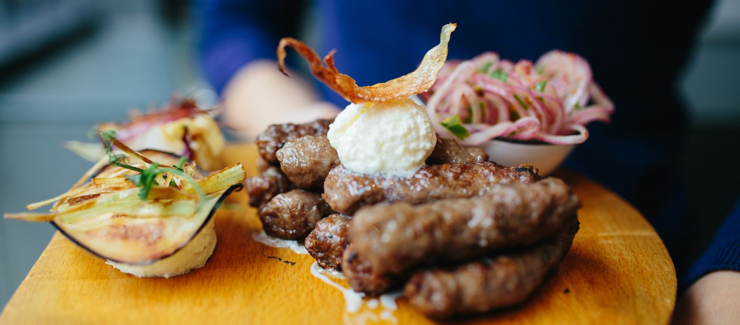 Plate of food, ćevapi, served on a wooden board. Grilled minced meat rolls, topped with a dollop of a white creamy substance and a crispy garnish. On the side, there are slices of what appear to be grilled onion and a small bowl of finely sliced red onions