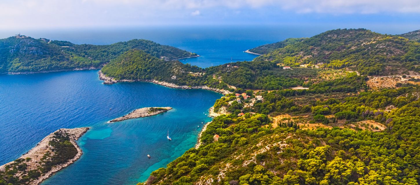 National park with lush forests, natural harbors, and clear waters. Bay with several small, green islands and peninsulas covered in dense vegetation. The water is a vibrant blue and turquoise, with a small sailboat visible in the bay. The landscape features rocky coastlines and a few buildings nestled among the trees.