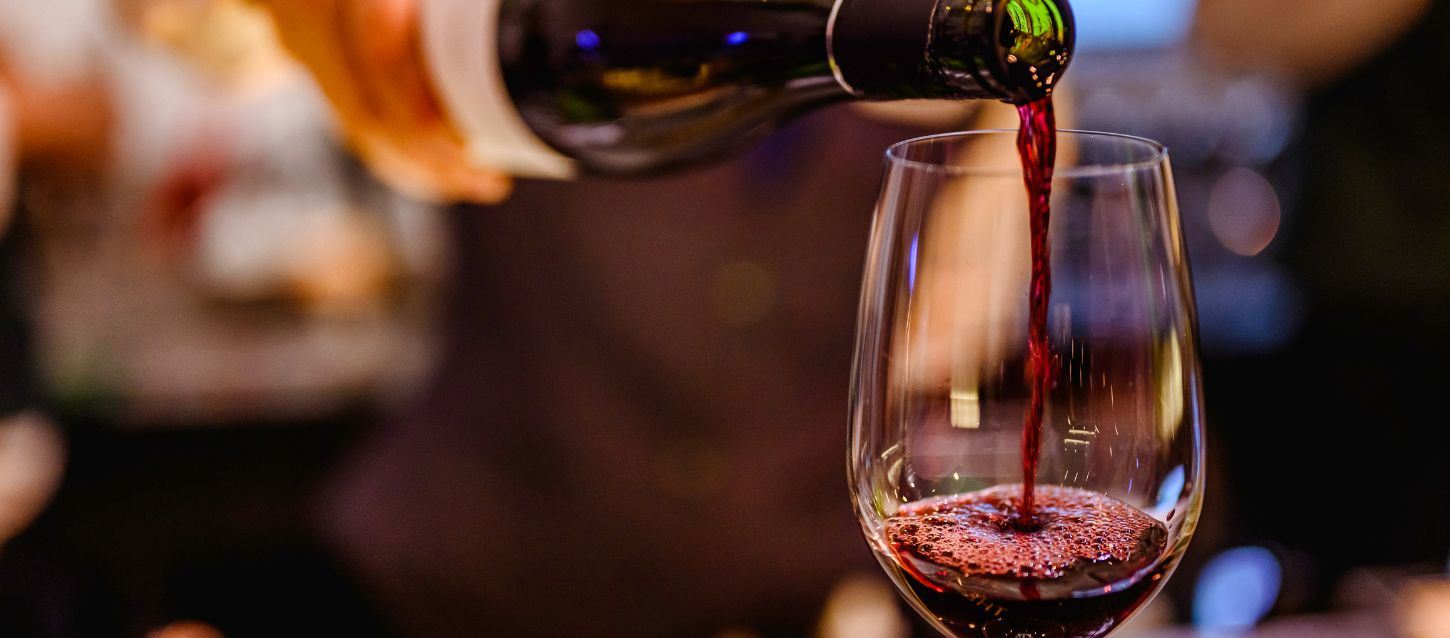 close-up shot of a bottle pouring red wine into a clear wine glass. The wine is a deep red color, and the glass is partially filled with the liquid, showing some bubbles and foam at the surface. The background is dark and out of focus, suggesting a low-light setting like a bar or restaurant.