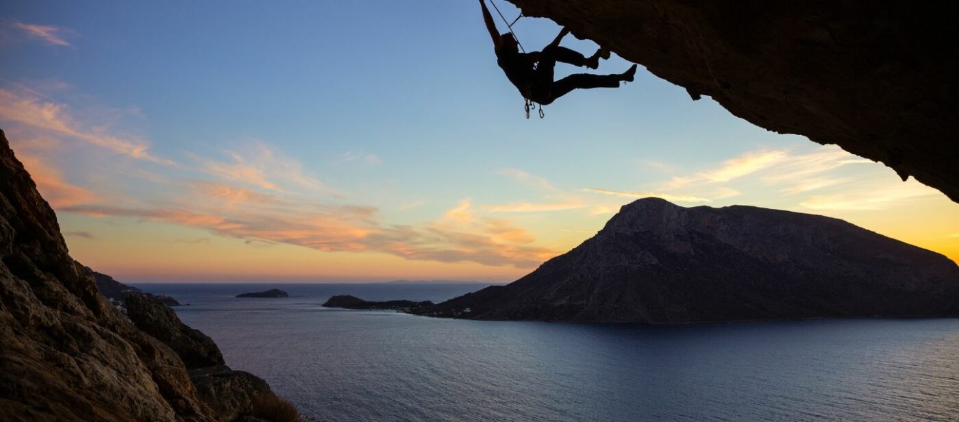 A view of the Acropolis of Lindos in Rhodes, Greece. The image shows ancient stone columns and a wall on a cliff overlooking the turquoise sea and a small bay. The sky is bright blue and clear.