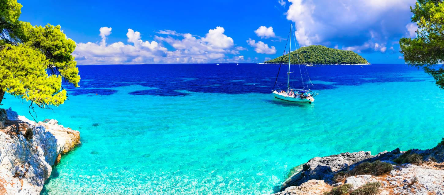 A sailboat anchored in a bay with incredibly clear, turquoise water, with a lush green island in the background under a blue sky with white clouds in Kastani.