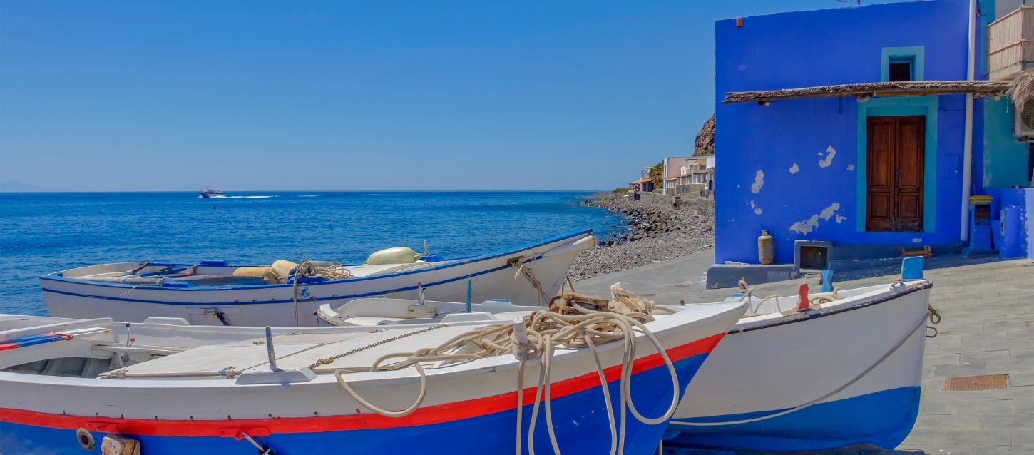 A picturesque seaside village with fishing boats on a rocky shore and a vibrant blue building under a clear sky. The scene is likely located on the island of Filicudi.