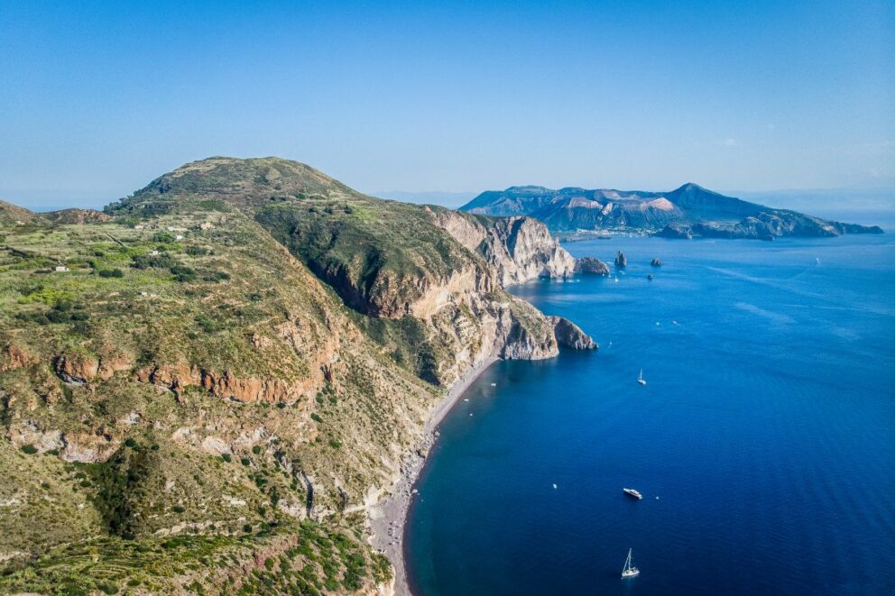 A scenic view of the volcanic island of Salina in the Aeolian Islands, Italy, with lush greenery and colorful flowers in the foreground. The island's twin peaks, Monte Fossa delle Felci and Monte dei Porri, rise from the sea under a partly cloudy sky. The image captures the island's natural beauty and serene atmosphere.
