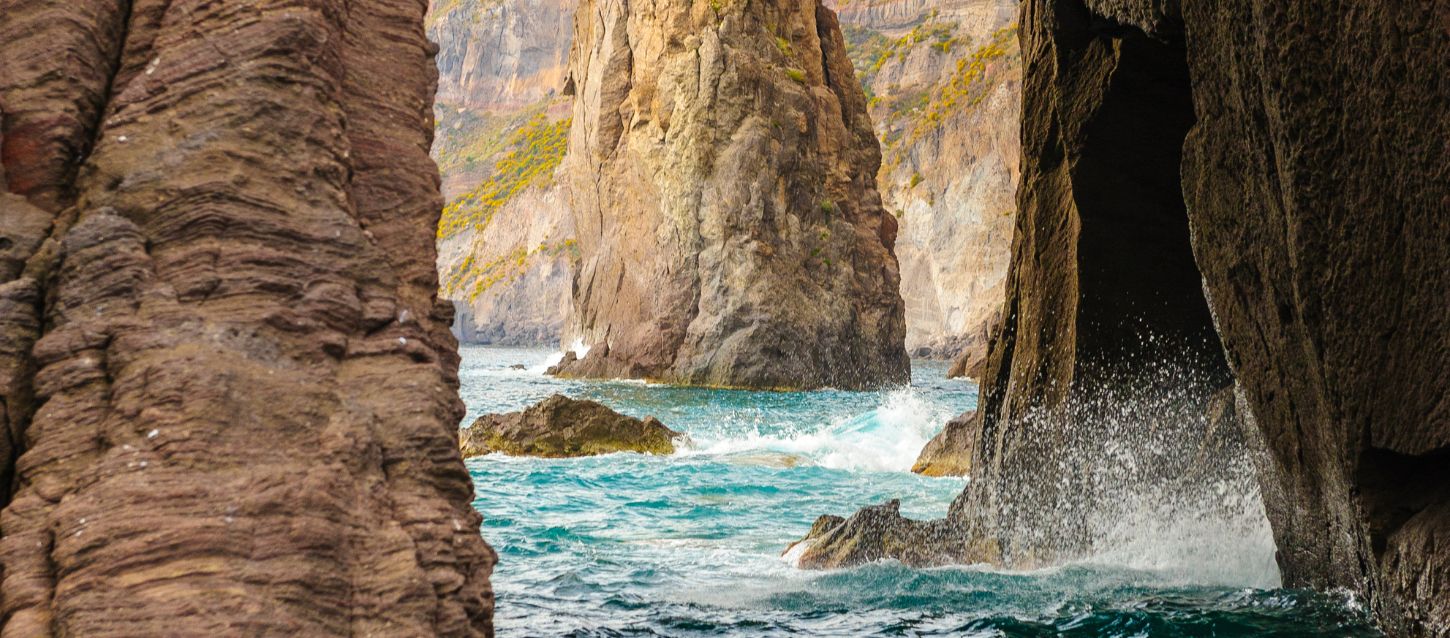 A dramatic view of a rocky coastline with waves crashing against sea stacks and cliffs, seen from within a cave or between two large rock formations. The water is a vibrant turquoise and deep blue.