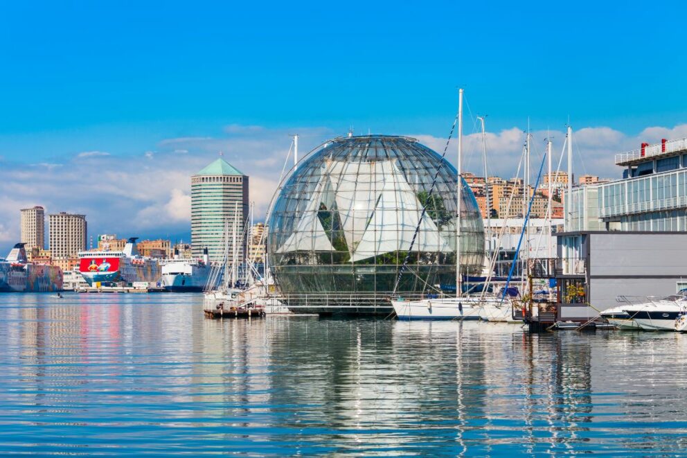 Modern waterfront view of Genoa, Italy, showing the glass Biosphere structure, moored sailboats, and the city skyline with colorful buildings and a cylindrical tower under a bright blue sky.