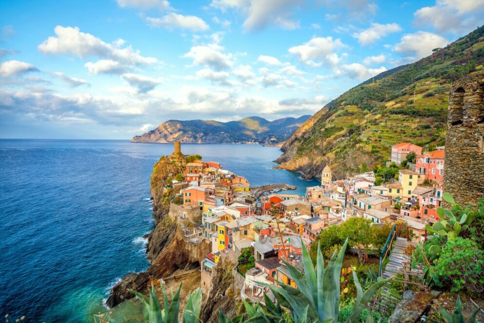 Colorful coastal village perched on steep cliffs overlooking the Mediterranean Sea in Vernazza, Cinque Terre, Italy, with terraced hillsides and blue sky filled with scattered clouds.