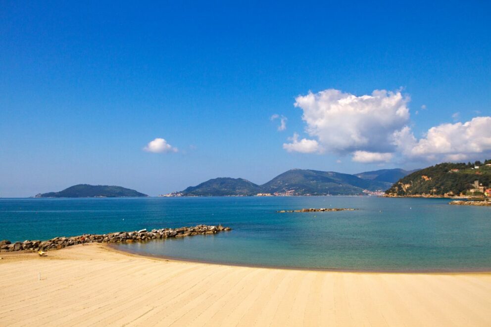 Peaceful sandy beach with calm turquoise water, distant green hills, and a few scattered clouds in the bright blue sky in Lerici, Italian Riviera.