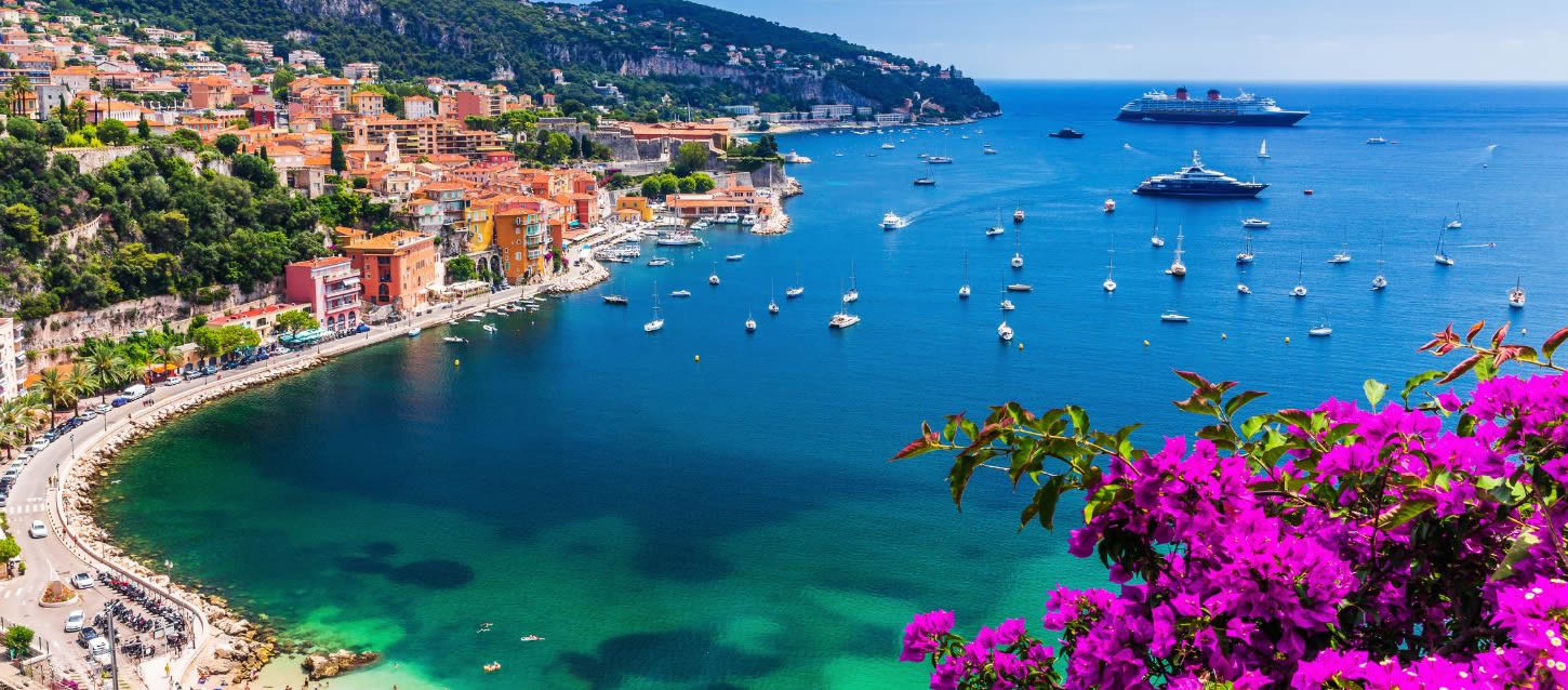 Panoramic view of the natural bay of Villefranche-sur-Mer with the promenade overlooking the turquoise sea with moored yachts and the flowering bougainvillea vegetation and coastline.
