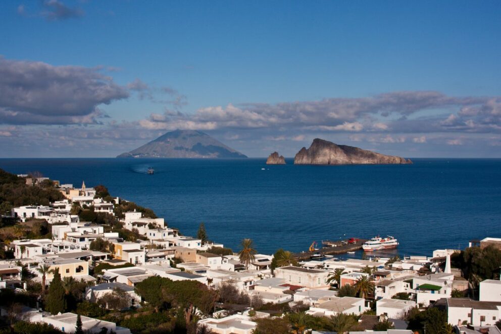 A scenic view of a coastal town with white buildings on a hillside, overlooking the Tyrrhenian Sea. In the distance, the volcanic island of Stromboli is visible, along with another smaller island, under a blue sky with scattered clouds.