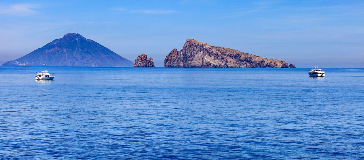 A scenic view of the sea with several islands in the distance, including the volcanic island of Stromboli, and two boats sailing on the water.