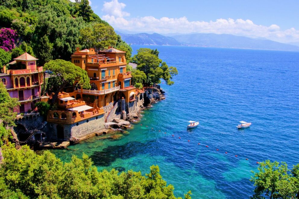Colorful villas built into a lush green hillside overlooking the turquoise waters of Portofino on the Italian Riviera, with small boats floating nearby.