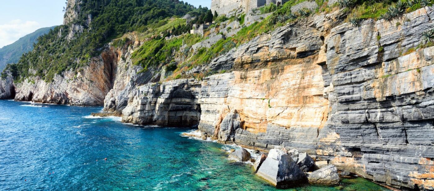 Rocky cliffs and turquoise waters near Portovenere, Italy, with lush green hills and ancient stone walls above the dramatic coastline.