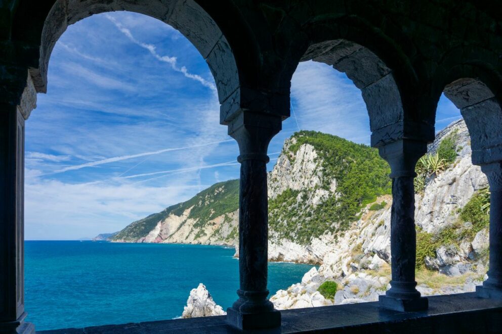 View of the rocky Ligurian coastline and turquoise sea through ancient stone arches in Portovenere, Italy, on a bright sunny day with wispy clouds above.
