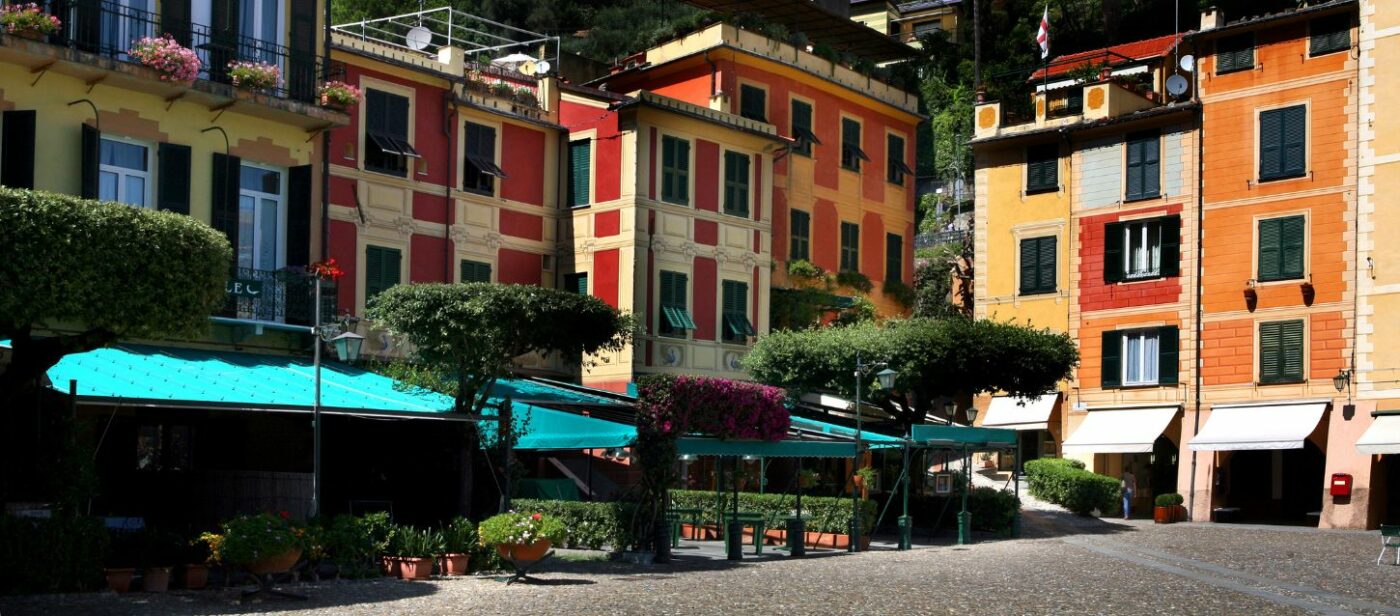 Charming town square in Portofino, Italy, with colorful pastel buildings, green awnings, manicured trees, and potted plants under bright sunlight.