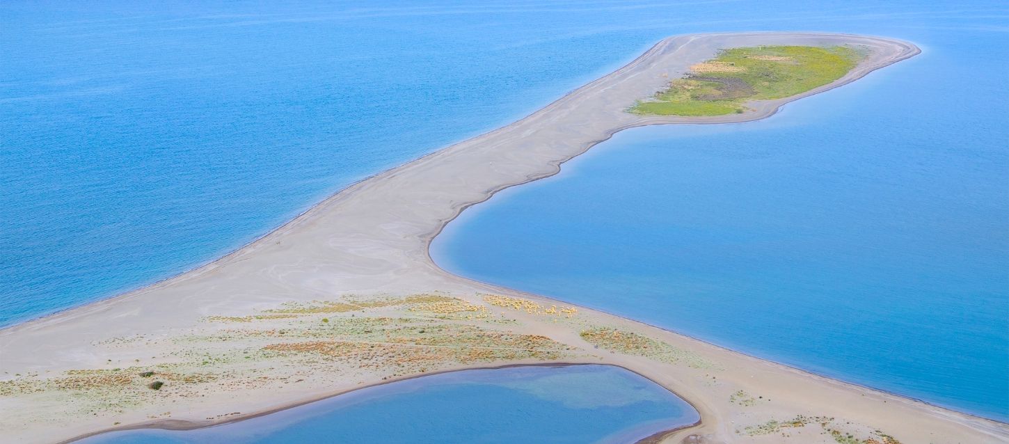 A long, curved sandbar extends into a body of blue water, with small patches of green vegetation on the land. The water is a mix of deep blue and lighter turquoise tones, especially in the small lagoon-like area formed by the curve of the sandbar
